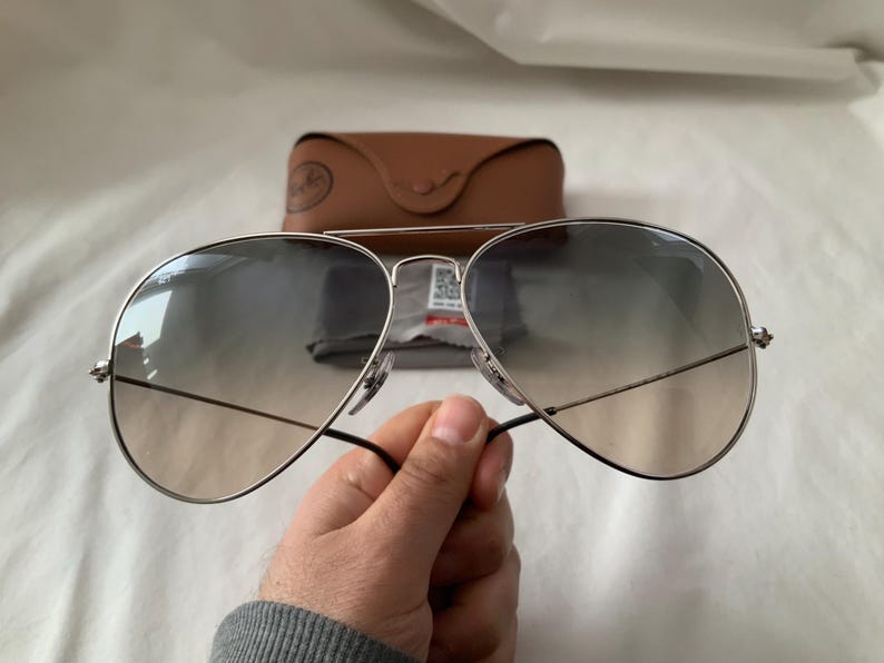 May include: Silver-framed aviator sunglasses with gradient lenses. The sunglasses have a double bridge and black temples. A brown leather case and cleaning cloth are in the background.