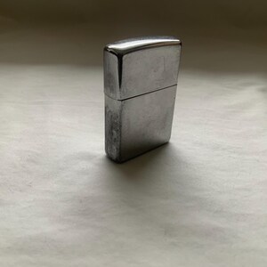 Puede incluir: Un encendedor de metal plateado con forma rectangular. El encendedor tiene una tapa con bisagras y un aspecto desgastado, lo que sugiere uso. La superficie muestra signos de desgaste.