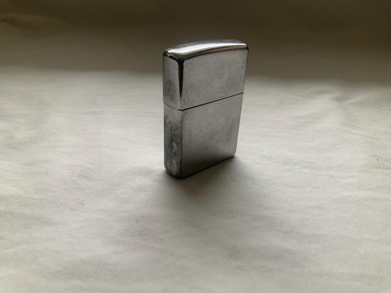 Puede incluir: Un encendedor de metal plateado con forma rectangular. El encendedor tiene una tapa con bisagras y un aspecto desgastado, lo que sugiere uso. La superficie muestra signos de desgaste.