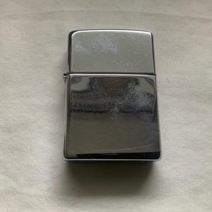 May include: A rectangular, silver-colored lighter with a hinged lid. The lighter has a shiny, reflective surface with visible scratches. The lighter is closed and sits on a white surface.