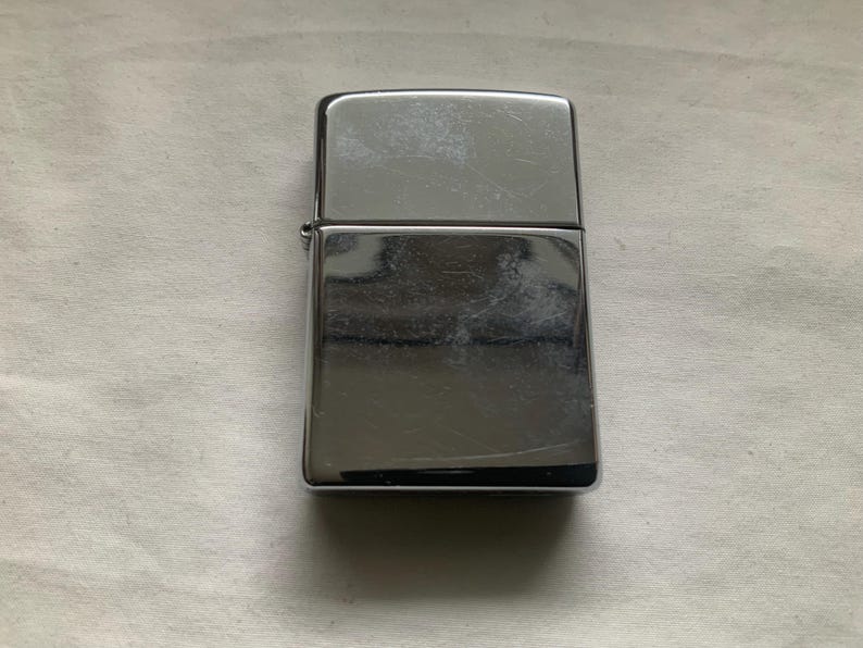 May include: A rectangular, silver-colored lighter with a hinged lid. The lighter has a shiny, reflective surface with visible scratches. The lighter is closed and sits on a white surface.