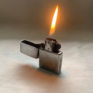 Puede incluir: Un encendedor de metal plateado con la tapa abierta y una llama encendida. La llama es amarilla y naranja, proyectando un brillo c&aacute;lido. El encendedor es rectangular y tiene una tapa con bisagras.