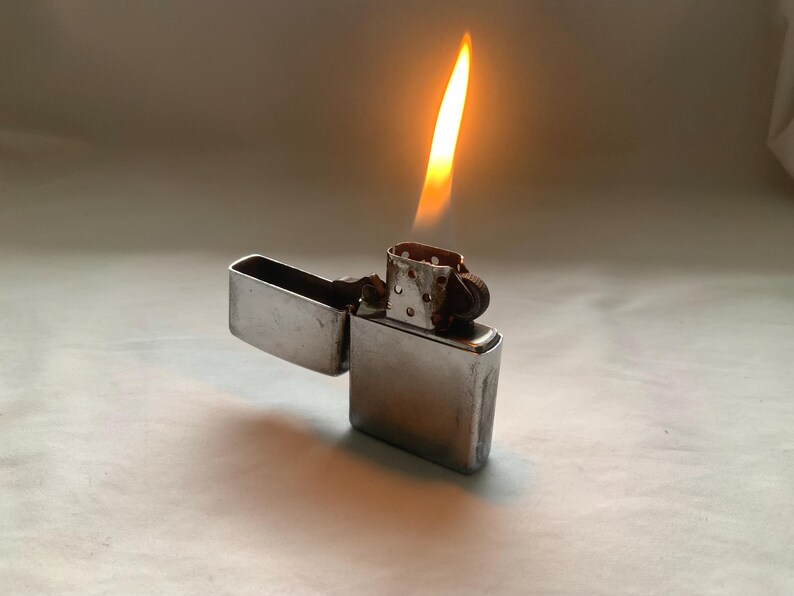 Puede incluir: Un encendedor de metal plateado con la tapa abierta y una llama encendida. La llama es amarilla y naranja, proyectando un brillo c&aacute;lido. El encendedor es rectangular y tiene una tapa con bisagras.