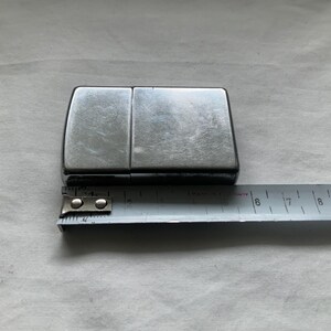 Puede incluir: Un encendedor rectangular de color plateado con tapa abisagrada. El encendedor se coloca junto a una cinta m&eacute;trica de metal, que muestra sus dimensiones. El encendedor parece ser de metal y tiene un acabado brillante.
