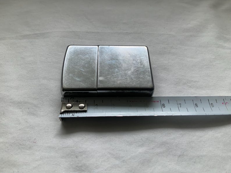 Puede incluir: Un encendedor rectangular de color plateado con tapa abisagrada. El encendedor se coloca junto a una cinta m&eacute;trica de metal, que muestra sus dimensiones. El encendedor parece ser de metal y tiene un acabado brillante.