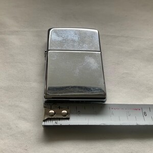 May include: A shiny, silver-colored lighter with a rectangular shape. The lighter is approximately 2 inches long. A measuring tape is placed next to the lighter.