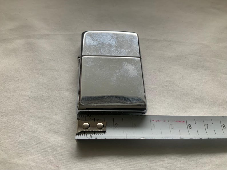 May include: A shiny, silver-colored lighter with a rectangular shape. The lighter is approximately 2 inches long. A measuring tape is placed next to the lighter.