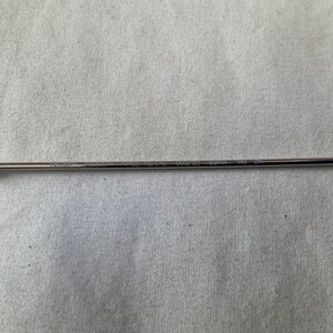 May include: A thin, silver-colored eyeglass arm with a black temple tip. The arm has etched text along its length. The background is a light gray fabric.