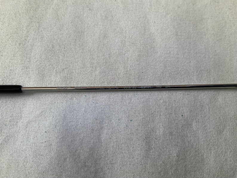 May include: A thin, silver-colored eyeglass arm with a black temple tip. The arm has etched text along its length. The background is a light gray fabric.