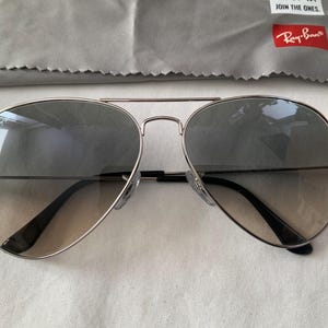 May include: Silver-framed aviator sunglasses with gradient lenses. The lenses transition from a darker shade at the top to a lighter tint towards the bottom. Includes a gray cleaning cloth and a red Ray-Ban logo.