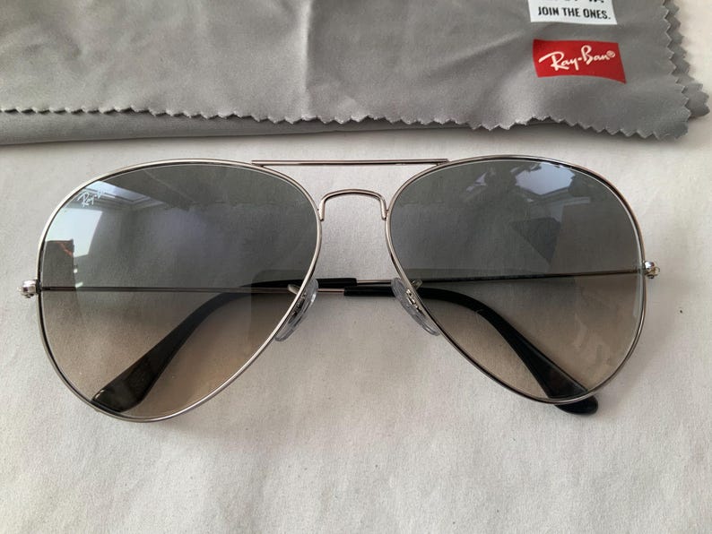 May include: Silver-framed aviator sunglasses with gradient lenses. The lenses transition from a darker shade at the top to a lighter tint towards the bottom. Includes a gray cleaning cloth and a red Ray-Ban logo.