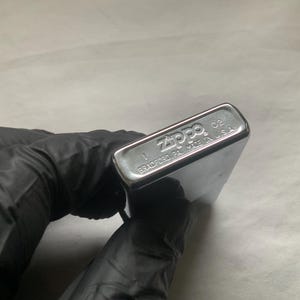May include: A silver Zippo lighter with the brand name and location of manufacture, Bradford, PA, Made in U.S.A., visible on the bottom. The lighter is held by a gloved hand.
