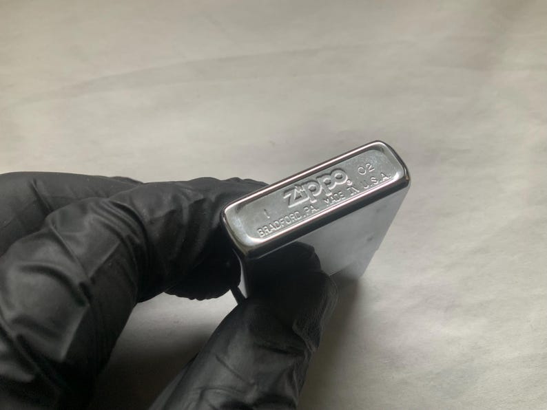 May include: A silver Zippo lighter with the brand name and location of manufacture, Bradford, PA, Made in U.S.A., visible on the bottom. The lighter is held by a gloved hand.