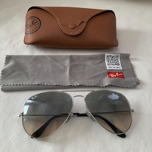 May include: Silver-framed aviator sunglasses with gradient lenses, a brown leather case, and a gray cleaning cloth. The case has a button closure and a logo. The cloth has a QR code and the text "JOIN THE ONES."