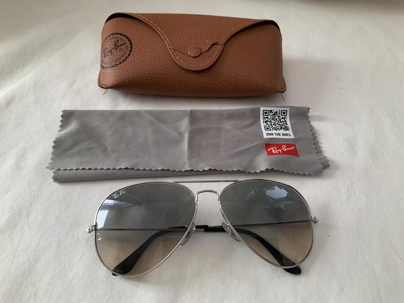 May include: Silver-framed aviator sunglasses with gradient lenses, a brown leather case, and a gray cleaning cloth. The case has a button closure and a logo. The cloth has a QR code and the text "JOIN THE ONES."