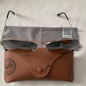 May include: Silver-framed sunglasses with dark lenses, a gray cleaning cloth, and a brown leather case. The case has a snap closure and a Ray-Ban logo. The cleaning cloth has a QR code and the text "JOIN THE ONES."