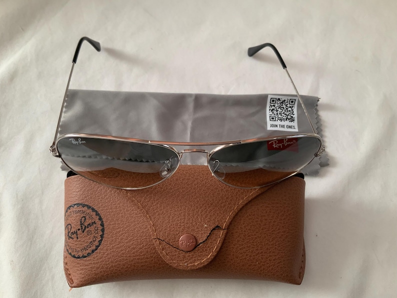 May include: Silver-framed sunglasses with dark lenses, a gray cleaning cloth, and a brown leather case. The case has a snap closure and a Ray-Ban logo. The cleaning cloth has a QR code and the text "JOIN THE ONES."