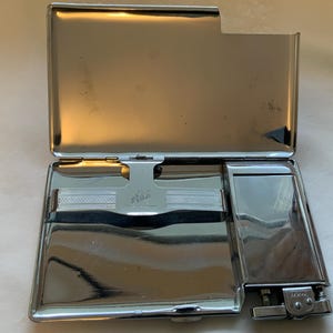 Puede incluir: Estuche de cigarrillos plateado brillante abierto con un encendedor incorporado. El estuche es rectangular con una tapa con bisagras y un compartimento para cigarrillos. El encendedor está adjunto al lado.