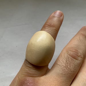 Puede incluir: Un anillo grande de madera de color claro, con forma ovalada y redondeada. El anillo se lleva en un dedo, mostrando su superficie lisa y pulida. El diseño del anillo es simple y natural, destacando la veta de la madera.