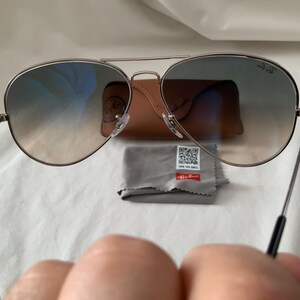 May include: Aviator sunglasses with gradient lenses, a silver frame, and black temples. The image includes a brown leather case, a gray cleaning cloth, and a small tag with text. A classic eyewear accessory.