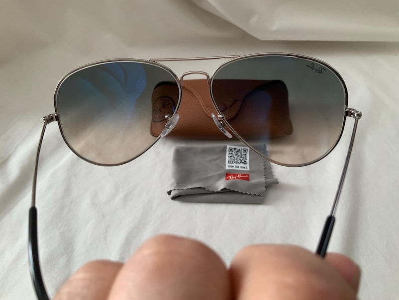 May include: Aviator sunglasses with gradient lenses, a silver frame, and black temples. The image includes a brown leather case, a gray cleaning cloth, and a small tag with text. A classic eyewear accessory.