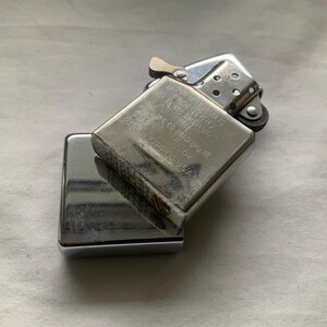 May include: A silver-colored Zippo lighter, with the top open, revealing the flint wheel and wick. The lighter is resting on top of its closed, rectangular base. The text on the lighter reads "Zippo, Bradford, PA. Made in USA."