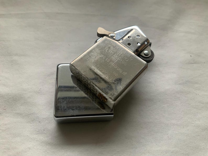 May include: A silver-colored Zippo lighter, with the top open, revealing the flint wheel and wick. The lighter is resting on top of its closed, rectangular base. The text on the lighter reads "Zippo, Bradford, PA. Made in USA."