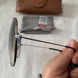 May include: Round sunglasses with a silver frame and gradient lenses. The temples are thin and extend to black earpieces. A brown case and cleaning cloth are in the background.