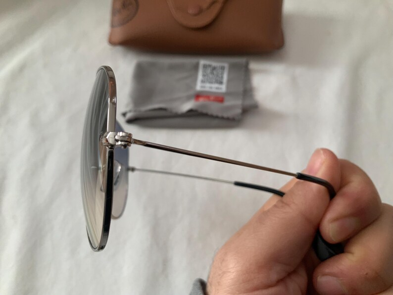 May include: Round sunglasses with a silver frame and gradient lenses. The temples are thin and extend to black earpieces. A brown case and cleaning cloth are in the background.