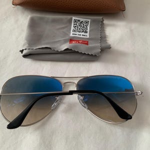May include: Aviator sunglasses with blue gradient lenses and silver frames. Includes a gray cleaning cloth with the text "JOIN THE ONES" and a brown case. The Ray-Ban logo is visible on the cloth.