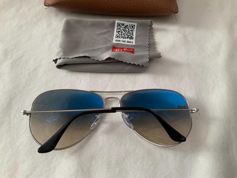 May include: Aviator sunglasses with blue gradient lenses and silver frames. Includes a gray cleaning cloth with the text "JOIN THE ONES" and a brown case. The Ray-Ban logo is visible on the cloth.