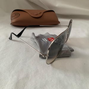 May include: Aviator-style sunglasses with silver frames and dark lenses, resting on a gray cleaning cloth. A brown leather case is in the background. The sunglasses have black temples and a classic design.