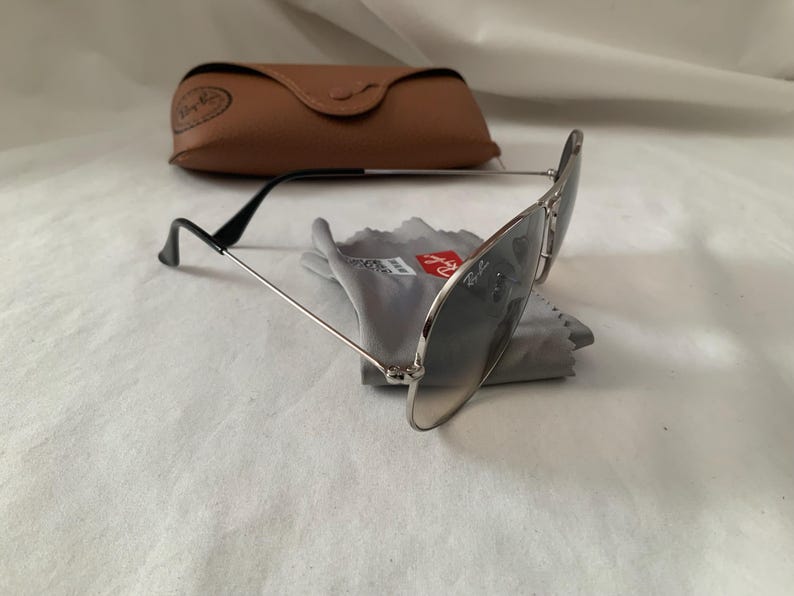 May include: Aviator-style sunglasses with silver frames and dark lenses, resting on a gray cleaning cloth. A brown leather case is in the background. The sunglasses have black temples and a classic design.