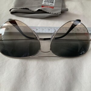 May include: Silver-framed sunglasses with gradient lenses. The sunglasses have a double bridge and black temples. A cleaning cloth and a ruler are also in the image. The sunglasses have a logo on the lens.