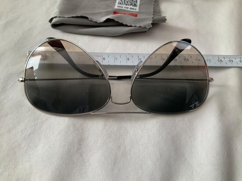 May include: Silver-framed sunglasses with gradient lenses. The sunglasses have a double bridge and black temples. A cleaning cloth and a ruler are also in the image. The sunglasses have a logo on the lens.