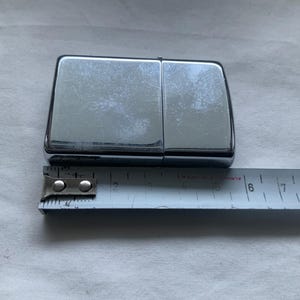 May include: A shiny, chrome-colored lighter with a rectangular shape. A metal measuring tape is placed next to the lighter, showing a measurement of approximately 2 inches. The lighter is on a white surface.
