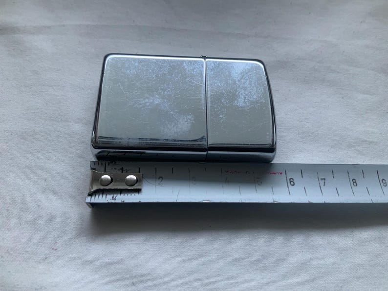 May include: A shiny, chrome-colored lighter with a rectangular shape. A metal measuring tape is placed next to the lighter, showing a measurement of approximately 2 inches. The lighter is on a white surface.