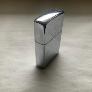 May include: A shiny, chrome-colored lighter. The rectangular lighter has a hinged lid and a smooth, reflective surface. The lighter is standing upright on a light-colored surface.