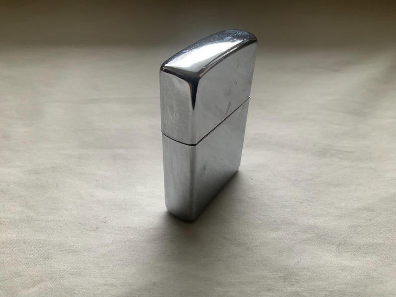 May include: A shiny, chrome-colored lighter. The rectangular lighter has a hinged lid and a smooth, reflective surface. The lighter is standing upright on a light-colored surface.