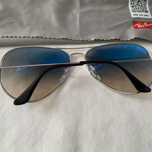 May include: Aviator sunglasses with gradient blue lenses and silver frames. The sunglasses have black temples and nose pads. A gray cleaning cloth with the Ray-Ban logo is in the background.