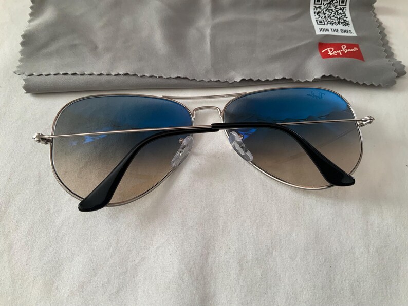 May include: Aviator sunglasses with gradient blue lenses and silver frames. The sunglasses have black temples and nose pads. A gray cleaning cloth with the Ray-Ban logo is in the background.