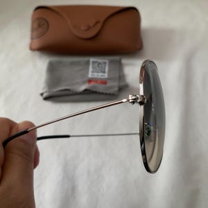 May include: Round sunglasses with silver frames and dark lenses are held in front of a white background. A brown leather case, a gray cleaning cloth, and a small tag are in the background.