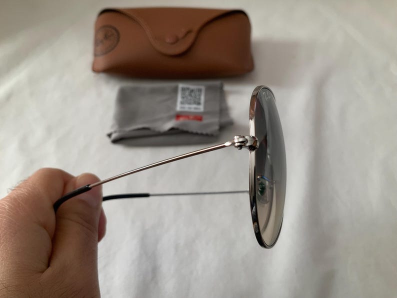 May include: Round sunglasses with silver frames and dark lenses are held in front of a white background. A brown leather case, a gray cleaning cloth, and a small tag are in the background.