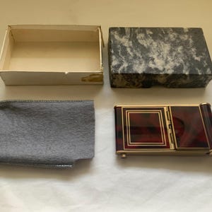 May include: A rectangular gold-tone metal case with a dark red and black tortoiseshell pattern, featuring a hinged lid and gold border accents. It includes a gray felt protective sleeve, a white cardboard box base, and a black and white marbled lid.
