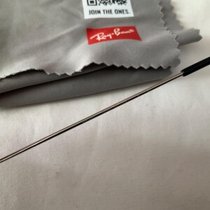 May include: Close-up of a pair of Ray-Ban sunglasses with a silver frame and black temple tips. A gray microfiber cleaning cloth with the Ray-Ban logo and the text "JOIN THE ONES" is in the background.