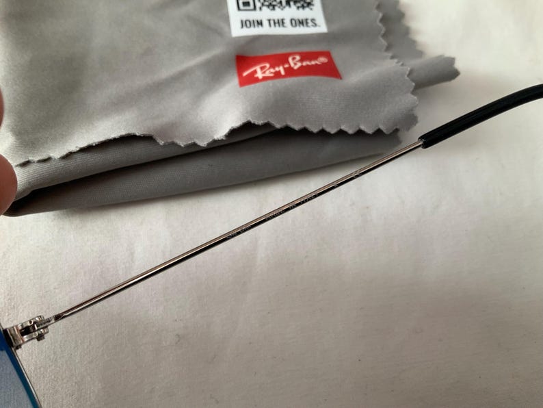 May include: Close-up of a pair of Ray-Ban sunglasses with a silver frame and black temple tips. A gray microfiber cleaning cloth with the Ray-Ban logo and the text "JOIN THE ONES" is in the background.