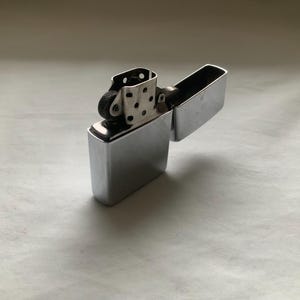 May include: A shiny, silver-colored lighter with a rectangular body and a hinged lid. The lighter is open, revealing the internal components, including the flint wheel and wick. The surface reflects light, creating highlights and shadows.