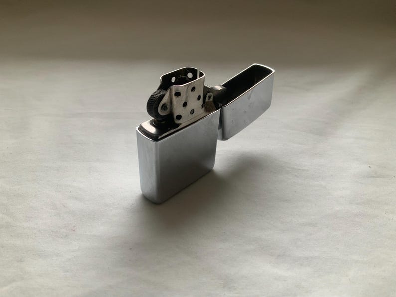 May include: A shiny, silver-colored lighter with a rectangular body and a hinged lid. The lighter is open, revealing the internal components, including the flint wheel and wick. The surface reflects light, creating highlights and shadows.