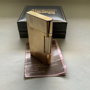 May include: A rectangular gold-tone metal lighter featuring a fine grid-patterned texture on its surface. The lighter is positioned in front of a black presentation box with the text Hadson printed on top and a paper instruction manual underneath.