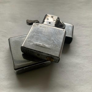 Puede incluir: Dos encendedores Zippo plateados apilados sobre una superficie clara. El encendedor superior est&aacute; abierto, revelando el interior. Los encendedores parecen usados, con algo de desgaste visible en el metal.